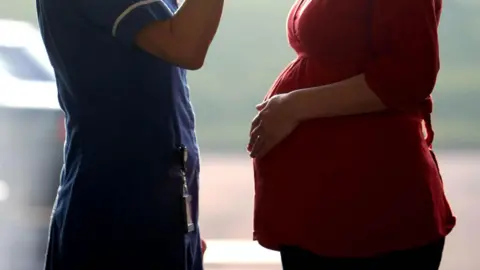 A nurse facing a pregnant woman. Their faces can't be seen. The nurse is wearing a blue uniform and the pregnant women is wearing a red dress.