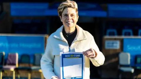 Southend United A woman standing in a football ground with a big smile and holding a sheet of paper