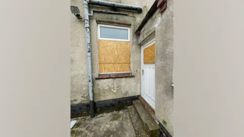 A rear entrance to a property. The window of the back door has been boarded up with ply wood. A window next to it has also been boarded up in a similar manner.