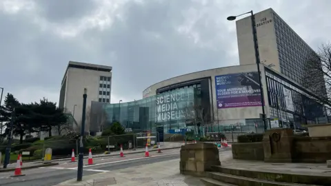 The front of the Science and Media Museum, which has a concrete facade behind a curved glass wall bearing the words Science + Media Museum in large white capital letters. On either side of the building are taller concrete tower blocks.