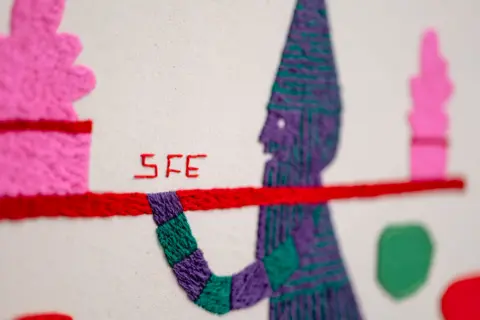 Stewart Francis Easton A close-up image of an embroidered character in purple and green, holding a red bar balancing pink and red shapes. 