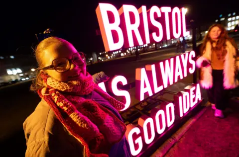 Two women stand next to a bright glowing neon sign that reads "Bristol is always a good idea"