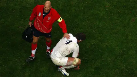 Getty Images Phil Pask, wearing shorts, a red top, and long socks, holding a black bag, with his arm on the shoulder of a player, crouched down, with a white top and white shorts on and has his back to the camera. 