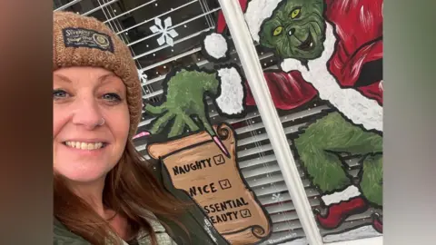 A woman with auburn hair is wearing a brown hat a green coat. She is taking a selfie in front of a window which has been painted. The drawing is of the grinch who is green, dressed in red and white Christmas clothing and a santa hat. He is holding a list in his hand that says "naughty" and "nice"