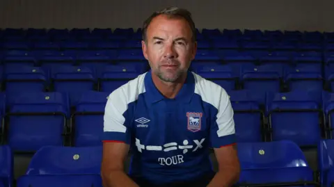 Matchday Images/ITFC Marcus Stewart sits in an empty football stadium on a blue seat and is pictured in a blue and white Ipswich Town shirt.