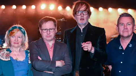 Sarah Jeynes/BBC Members of Pulp look at the camera. A woman in blue, with a green headband, looks to one side. A man with black-rimmed glasses and a jacket stands with his arms folded. Jarvis Cocker points directly at the camera and looks surprised. Another man stands to the right, wearing a blue short-sleeved shirt. Yellow spotlights are lit in the background.