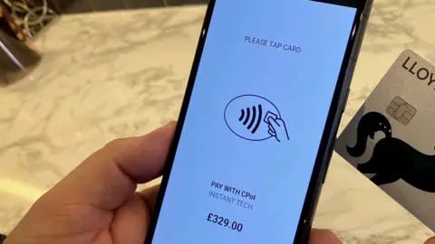 A mobile phone held in one hand showing an instruction to tap a payment card with the other hand holding a payment card