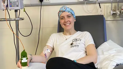 Vicky Green smiles as she sits in a chair receiving chemo treatment. She is holding a knitted green cactus toy. She is wearing a blue patterned head scarf.