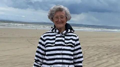 Family handout Joyce Taggart is smiling whilst wearing a black and white striped coat with neutral trousers. She is standing on the beach.