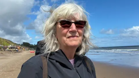 An older woman with shoulder length grey hair looks at the camera. She wears sunglasses and a black waterproof jacket. Behind her, a view of a beach and sea can be seen. 
