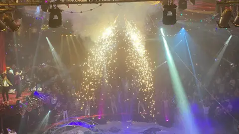 The inside of a circus with an audience sat in a semi circle around a stage. Pyrotechnics above the stage are producing bright gold sparks.