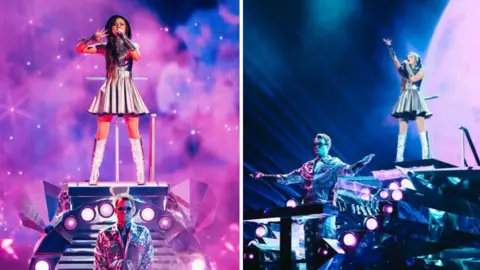 Sarah Louise Bennett / EBU A composite image of Emmy in a silver dress, stood on top of a stage, below her sits a man wearing sunglasses in a silver blazer