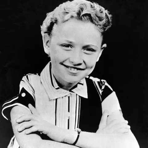 Getty Images/Michael Ochs Archives A black and white photo of Dolly as a child. She is sitting at a desk smiling and her hair is very short and curly. She wears a conservative black and white striped short sleeved shirt buttoned up to her neck.