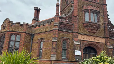 Clare Worden/BBC The ornate brick, turreted Carnegie Library on London Road in King's Lynn. The entrance has a tower on top of it, with the building lined with small trees, flowers and iron railings