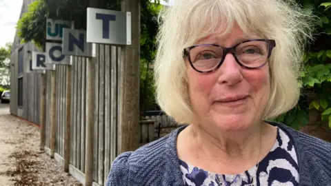 Venetia Young, who has a blonde bob and wears tortoiseshell-rimmed glasses, standing in front of a fence.