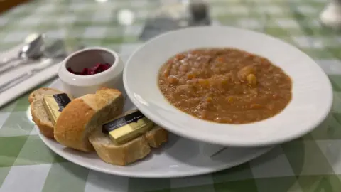 Jonny Humphries/BBC A white bowl containing a meat, potato and carrot stew on a plate with three slices of white crusty bread and butter and a small bowl of red cabbage resting on a green and white chequered table cloth. 