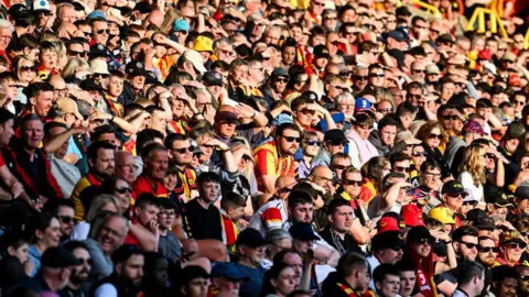 SNS Partick Thistle fans, mostly wearing the club's red and yellow striped top, sit in the stand of a football ground watching their team on a sunny day