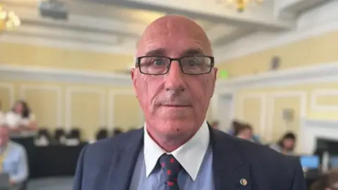 Nigel Hamilton. He is bald with blue eyes, wearing glasses, a dark grey blazer, light blue shirt with white collar, and black and red tie.