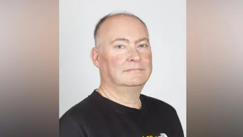 West Suffolk Council Roger Dicker wearing a black T-shirt standing in front of a white background looking at the camera