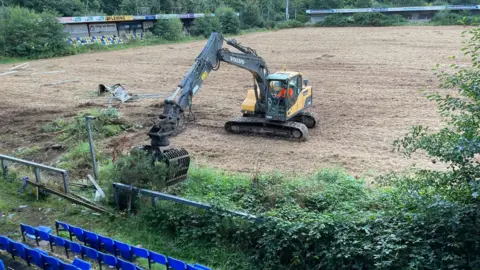 Andrew Russell A large yellow and black digger stands in the middle of a large pitch. 
Grassy bushes are near the seated stand of blue seats. 