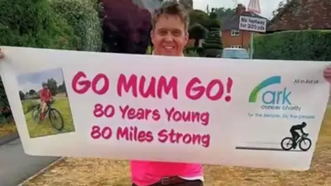 A family member smiling at the camera and holding a big sign which reads "Go Mum GO! 80 Years Young, 80 Miles Strong" Her son Alex is wearing green trousers and a pink t-shirt