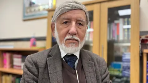 Abdul, a man with short white hair and white beard. He is wearing a checked jacket and a blue tie.