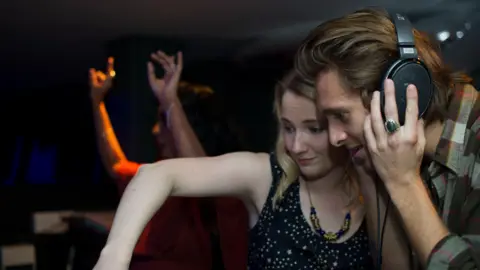 Pretty Ugly Lynne Johnston and Paolo Nutini behind the decks at a club night - he has on headphones and is wearing a chequed  shirt, while she has on a black dress.