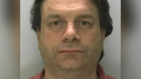 Richards looks at the camera in a blank expression in a custody picture. He is against a grey background, and has large grey eyebrows and dark hair. 