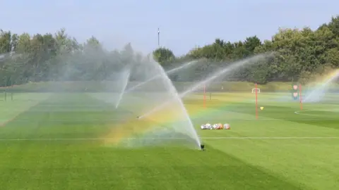 Getty Images A green football field with several water sprinklers spraying water in the air. 