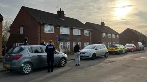 Louise Parry/BBC Police officers and members of the public stand on a residential street with cars, which includes a police vehicle, parked along the pavement.