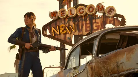 Prime Video Ella Purnell, dressed as character Lucy, stands next to a rusted, ruined car in front of a battered "Welcome to New Vegas" sign. She's holding a rifle as she looks off to the side, as if she's just spotted a sudden threat.