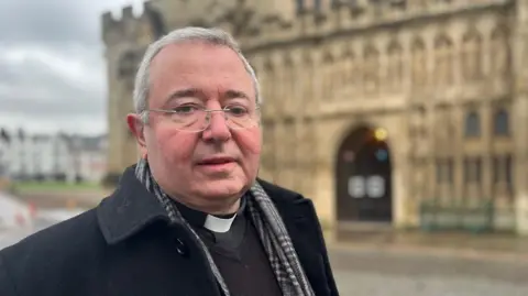 The Dean of Exeter, the Very Reverend Jonathan Greener, wearing a scarf and a black coat and standing in Cathedral Green with the cathedral in a blurred background.