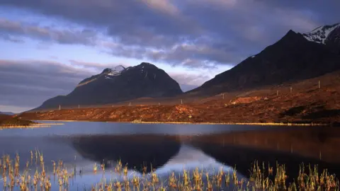 Towering mountains rise above an area of moorland and lochs under a cloudy sky. The picture shows Liathach and part of Beinn Eighe NNR.