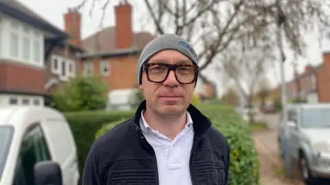 Nick Goodess is pictured wearing a grey beanie hat, glasses, a white shirt and black zipped jumper. Cars and houses can be seen blurred in the background.