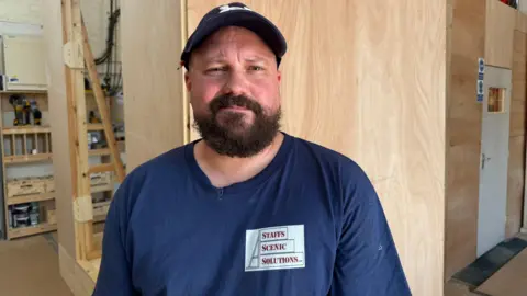 A man with a beard, wearing a hat, a dark blue T-shirt reading "Staffs scenic solutions", pictured from the shoulders up in front of a wooden wall with shelving.