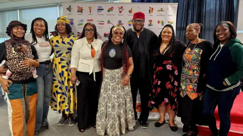 BAFA Media A group of nine people, all from an African-Caribbeanbackground, stand smiling in front of a display board which has the names and flag style maps of various African countries on it. Some of the group wear traditional African clothing.