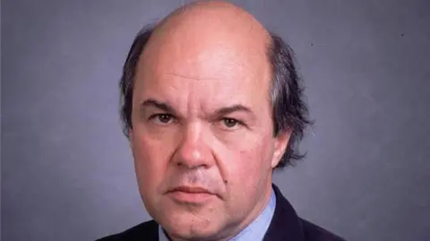 Portrait photo of a man with balding black hair. He is looking at the camera against a grey background, and wearing a black suit and blue shirt.