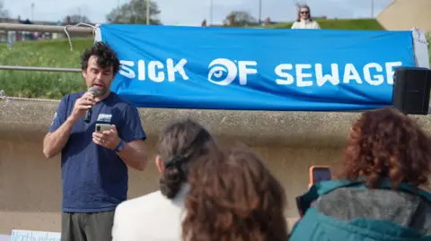 BBC/Stuart Prandle Nick Jones speaking into a microphone while looking down at his phone. He has dark, curly, short hair and is wearing a navy blue t-shirt and grey trousers. He is standing in front of a bright blue sign which reads 'sick of sewage'. The back of three people's heads are in the foreground of the photo, facing Jones.