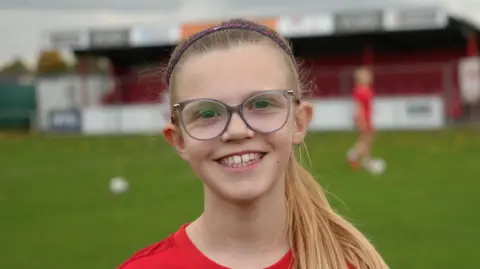 Shaun Whitmore/BBC Ruby smiles at the camera while standing on a football pitch in her red football shirt. She has long blonde hair that has been tied up with a purple headband and glasses.