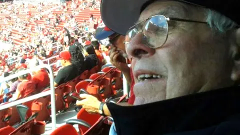 Cull family A picture of Raphael Cull in black hat and jacket and glasses, in the stands at a football match with crowd in background 