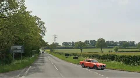 Google The B4208 road on a sunny day. The road is lined on one side with hedgerows and the other with trees. A car is visible in the distance, with another car in the foreground turning off the road at a junction.