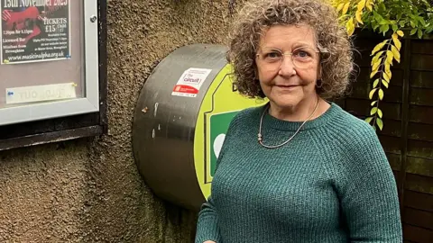 Lillian Berry is standing next to a box containing a defibrillator that is on the pebble-dashed wall of a building. The box is circular, with the curved edge silver-coloured and the flat front green and yellow. The sign and logo are obscured by Ms Berry, who is wearing a sea green jumper, silver chain necklace and round glasses. She has chin length curly hair and is tentatively smiling.