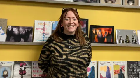 A woman with long brown hair standing in front of artwork sold in her shop. She is smiling and wearing a gold and black patterned jumper, orange hoop earrings and  glasses on her head.