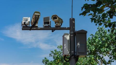 Two traffic enforcement cameras attached to a pole against a blue sky backdrop with some trees visible