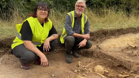 BBC/Rare TV Trish Biers and Oscar Aldred sitting in a pit which has revealed human remains. Trish is on the left and is wearing black trousers and a black T-shirt with a hi-vis yellow jacket over it. Oscar is on the right and is wearing dark trousers and a grey shirt with a hi-vis yellow waistcoat over it. In front of them is sandy soil and human remains including a spine and skull are emerging from the soil. 