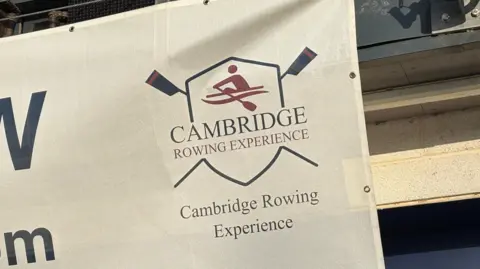 Ben Schofield/BBC The corner of a white banner, which is hung on the outside of a rowing club's boathouse. This part of the banner shows a logo with the words "Cambridge Rowing Experience" written underneath a simple drawing of a single rower in a boat, pulling a stroke on their oars. The rower image is set inside a shield, behind which are two crossed oars. Below the logo is a repeat of the words "Cambridge Rowing Experience".  