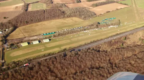 Andy Amor Aerial view of Popham Airfield and the A303 dual carriageway. There are two grass runways, green hangars and a series of other buildings surrounded by woodland.