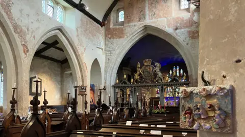 A view of the inside of the church - pews going towards an ornate altar with stained glass. Medieval archways with the remains of medieval paintwork in light red. To the right, the camera angle captures a small hanging nativity.