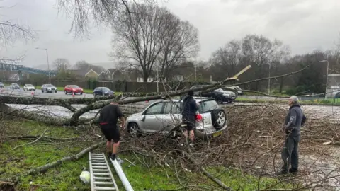 Danny Mac A tree falls on a car a silver car. There are three men who are trying to get the tree and debris of the car.