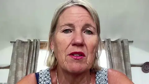 A blonde woman wearing pink lipstick looks into the camera from her home. You can see her curtains in the background. 